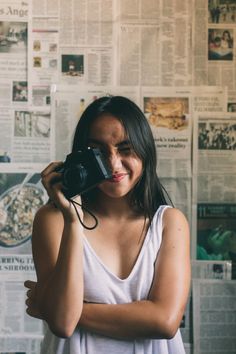 a woman holding a camera up to her face in front of a wall covered with newspapers