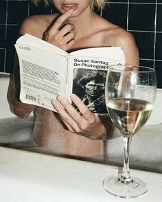 a woman sitting in a bathtub reading a book and holding a glass of wine