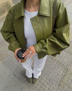 The outfit features an olive green leather jacket with a broad collar and front snap button detailing. It is layered over a crisp white tee, complemented by white pants. The ensemble is accessorized with a chain strap bag and green loafers. The person is holding a coffee cup, and their nails are manicured in a light pink shade, with a noticeable diamond ring on display. Scarlet Sage, Ideas Of Outfits, Outfit Ideas Stylish, Style Vert, Ladies Short Jackets, Ootd Women, Green Leather Jackets, Cropped Coat, Neutral Outfits