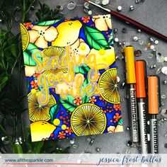 an open notebook with some markers and pens on the table next to it are lemons, leaves, and flowers