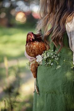 a woman in an apron holding a chicken and some flowers on her left arm with long hair