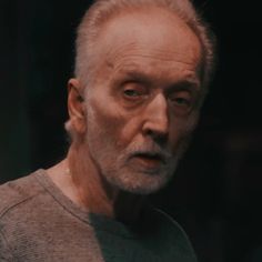 an older man with white hair and beards stares at the camera while standing in front of a dark background