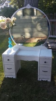 a white vanity table with drawers and a round mirror on the top, in front of a house