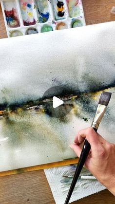 a person holding a paintbrush in front of a painting on a table with watercolors