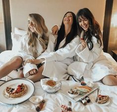 three women in robes sitting on a bed with food and drinks around them, smiling at the camera