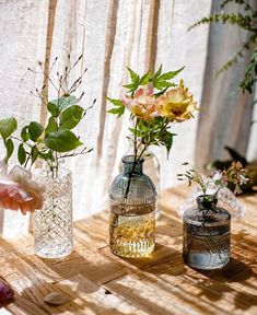 three vases filled with flowers sitting on top of a wooden table next to a curtain