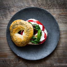 a bagel sandwich with tomatoes, cheese and spinach on a black plate sitting on a wooden table