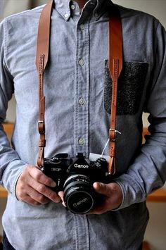 a man is holding a camera in his right hand and wearing a brown leather shoulder strap