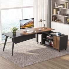 a desk with a computer on it in front of a window and bookshelf