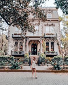 a woman standing in front of a large house with lots of plants on the sidewalk