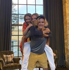 three girls are hugging each other in front of a large window with two chairs behind them