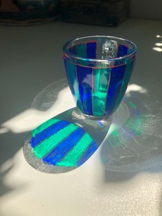 a blue and green glass cup sitting on top of a white table next to a shadow