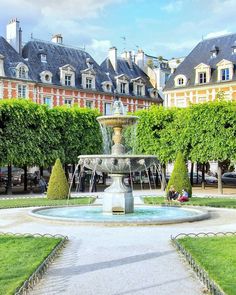 a fountain in the middle of a courtyard surrounded by trees and buildings with people sitting on benches