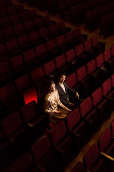 two people sitting in the middle of an empty auditorium with rows of red velvet seats