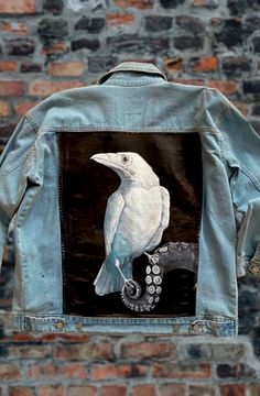 a denim jacket with a white bird painted on the front and back, hanging against a brick wall