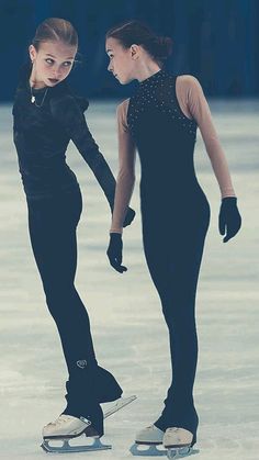 two women skating on an ice rink wearing black outfits and gloves, one is holding the other's hand