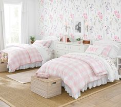 two beds in a room with floral wallpaper and pink bedding, one has a basket on the floor