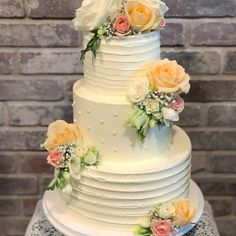 a three tiered white cake with flowers on the top and sides, against a brick wall