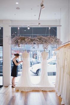 a woman looking at wedding dresses on display in a store window with flowers hanging from the windows