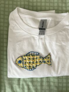 a white t - shirt with a fish embroidered on the front and back, sitting on a bed