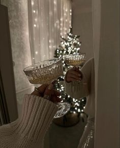 a woman holding two wine glasses in front of a christmas tree with lights on it