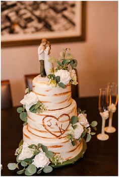 a wedding cake decorated with white flowers and greenery on a table next to wine glasses