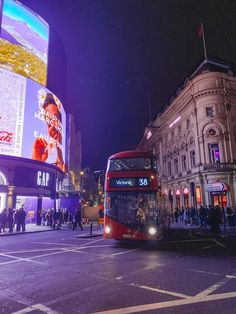 a red double decker bus driving down a street next to tall buildings with advertisements on it