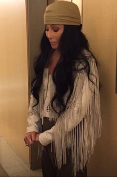 a woman with long black hair wearing a white shirt and fringed shawl standing next to a wall