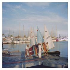 two men working on a sailboat in the water with other boats behind them at a dock