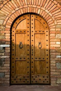 an arched wooden door with metal knobs on the side of a brick wall in front of a doorway