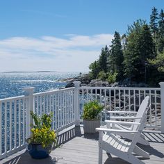 two white lawn chairs sitting on top of a wooden deck next to the ocean and trees