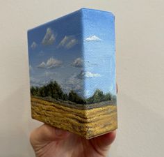 a hand is holding up a small box with a landscape painted on the front and sides