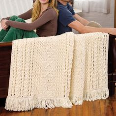 a man and woman sitting on top of a wooden bench next to a white crocheted blanket