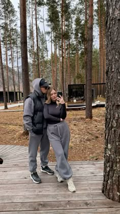 a man and woman standing on a wooden deck in front of pine trees with their arms around each other