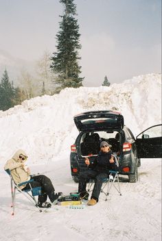 two people sitting in chairs next to a car on the snow covered ground with skis