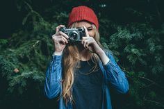 a woman holding up a camera to take a photo in front of some pine trees