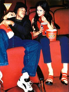 two people sitting on red seats in front of each other eating popcorn and drinking soda