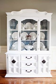 a white china cabinet with glass doors and plates on it's front, in the middle of a room