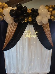 black and gold balloons are hanging from the ceiling in front of a white backdrop that reads let's celebrate