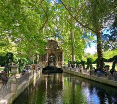 people sitting on benches near the water in a park with many trees and bushes around it