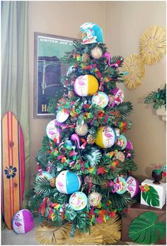 a colorful christmas tree decorated with beach balls and palm leaves, sits in front of a surfboard