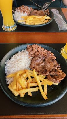 two plates filled with meat, rice and french fries on a table next to a glass of orange juice