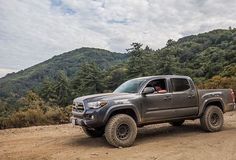 a gray truck driving down a dirt road next to a lush green forest covered hillside