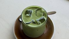 a green drink with ice cubes in it on a saucer next to a spoon