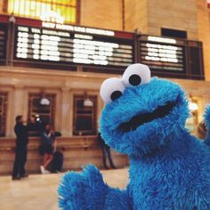 a blue stuffed animal sitting in front of a building with people standing around it and looking at the camera