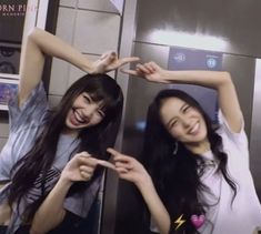 two young women standing next to each other in front of a metal wall holding their hands up