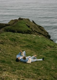 a man and woman laying on top of a lush green hillside next to the ocean