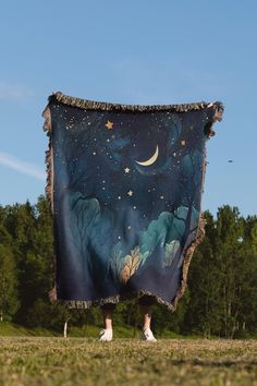 a person standing in the grass holding up a blanket with trees and stars on it