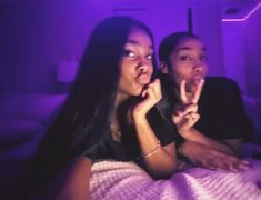 two people sitting on a bed with purple light in the background and one person holding something up to her mouth