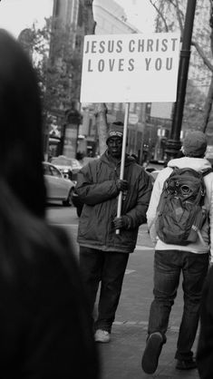 black and white photograph of two men walking down the street holding a sign that says jesus christ loves you